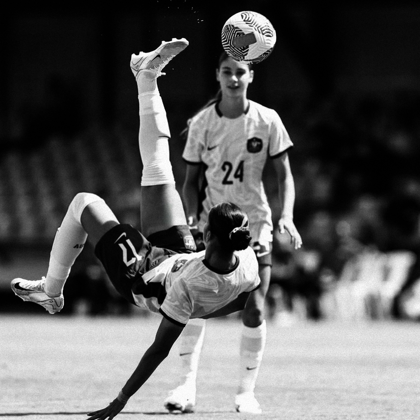 Black and white soccer scene: a player on the ground performs a bicycle kick while another player watches, ball hovering near the baller's head.