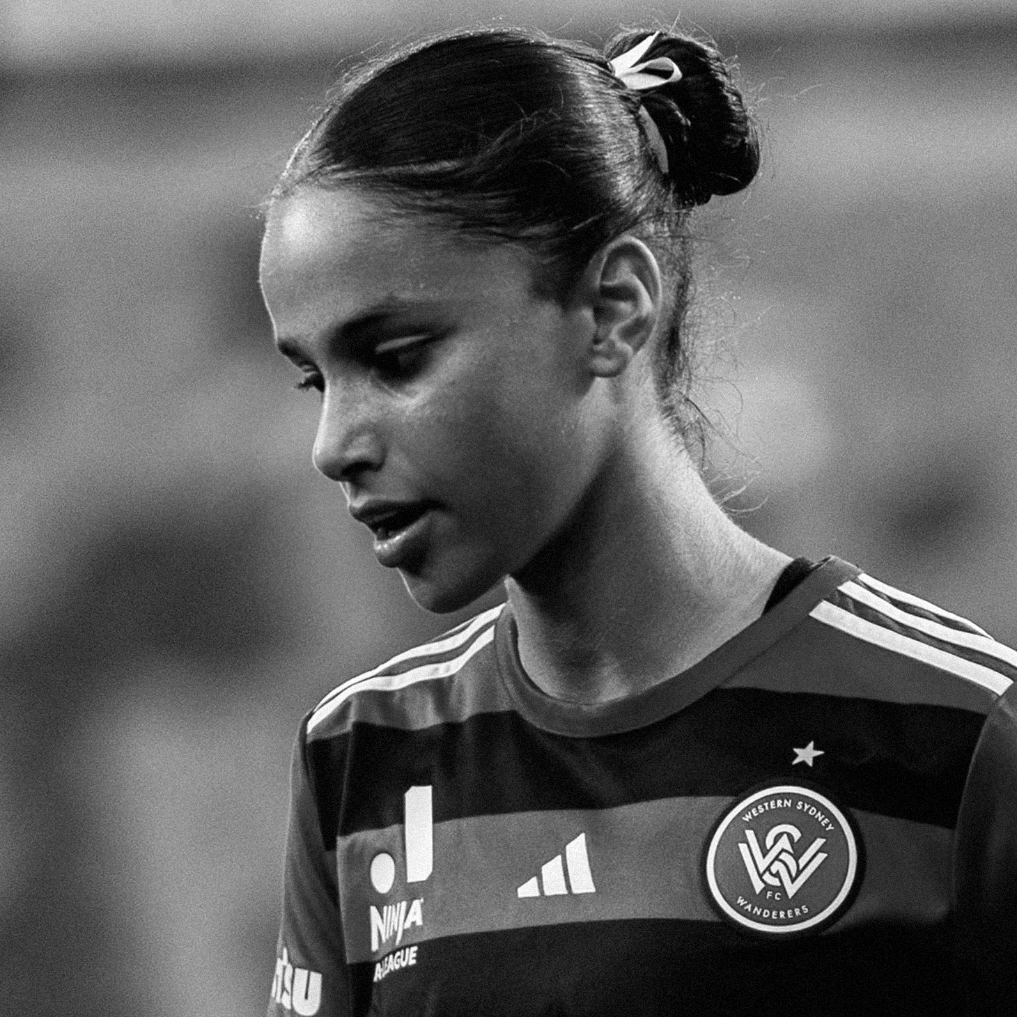 Female soccer player in a Wanderers jersey looking down, hair in a bun, focused on the field.